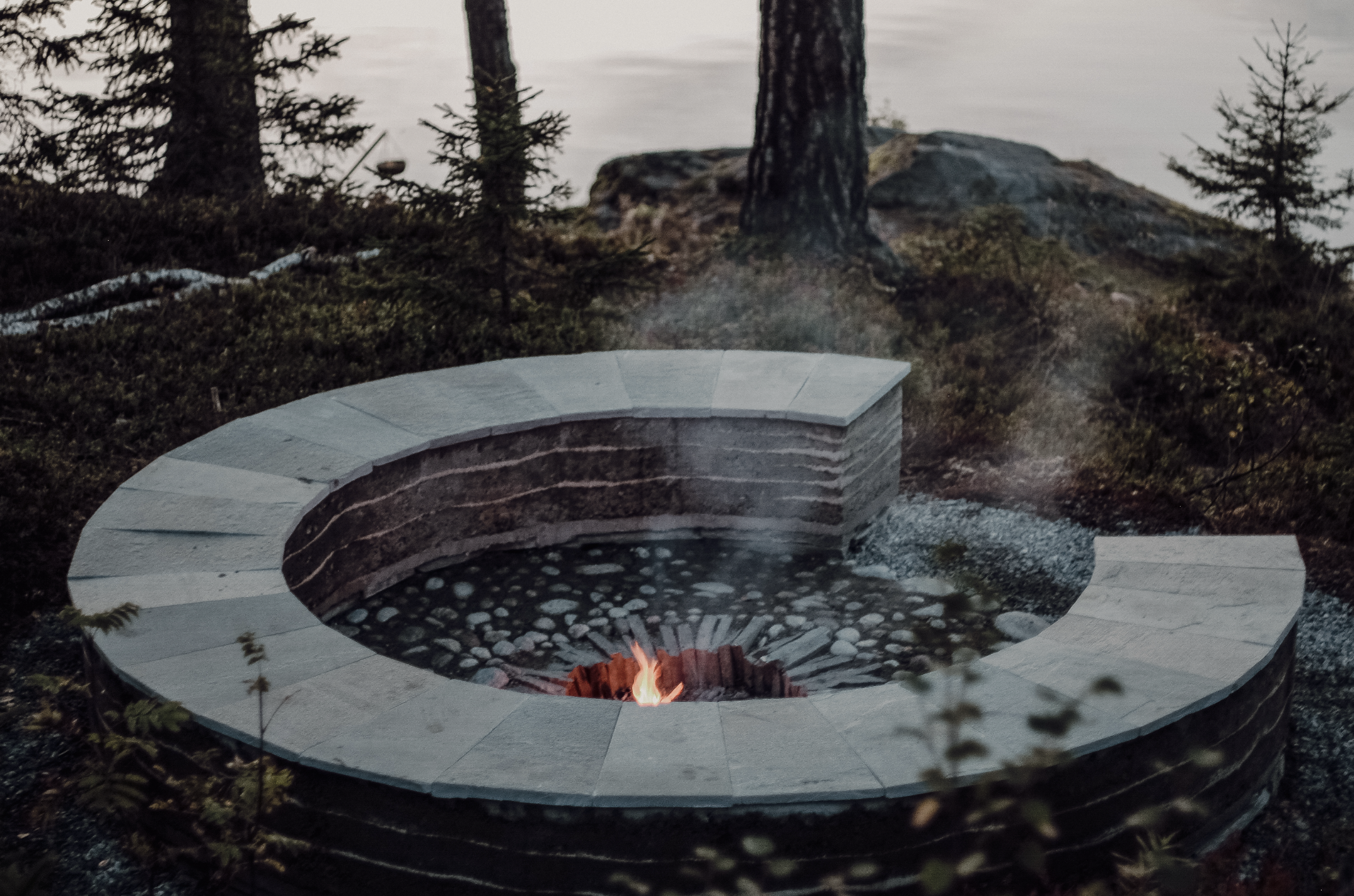 Sirkulær stampetjordkonstruksjon ved et vann hvor man kan sitte rundt et bål. Foto.